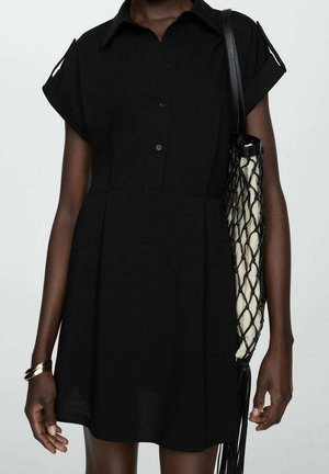 Femme portant une robe noire à manches courtes avec épaules découpées, tenant un sac fourre-tout noir en résille avec une pochette intérieure blanche, et un bracelet doré au poignet.