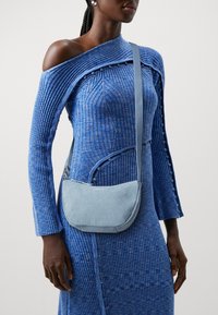 Žena v modrých žebrovaných úpletových šatech s jedním ramenem a světle modrou denimovou taškou Levi's přes rameno s nastavitelným popruhem.