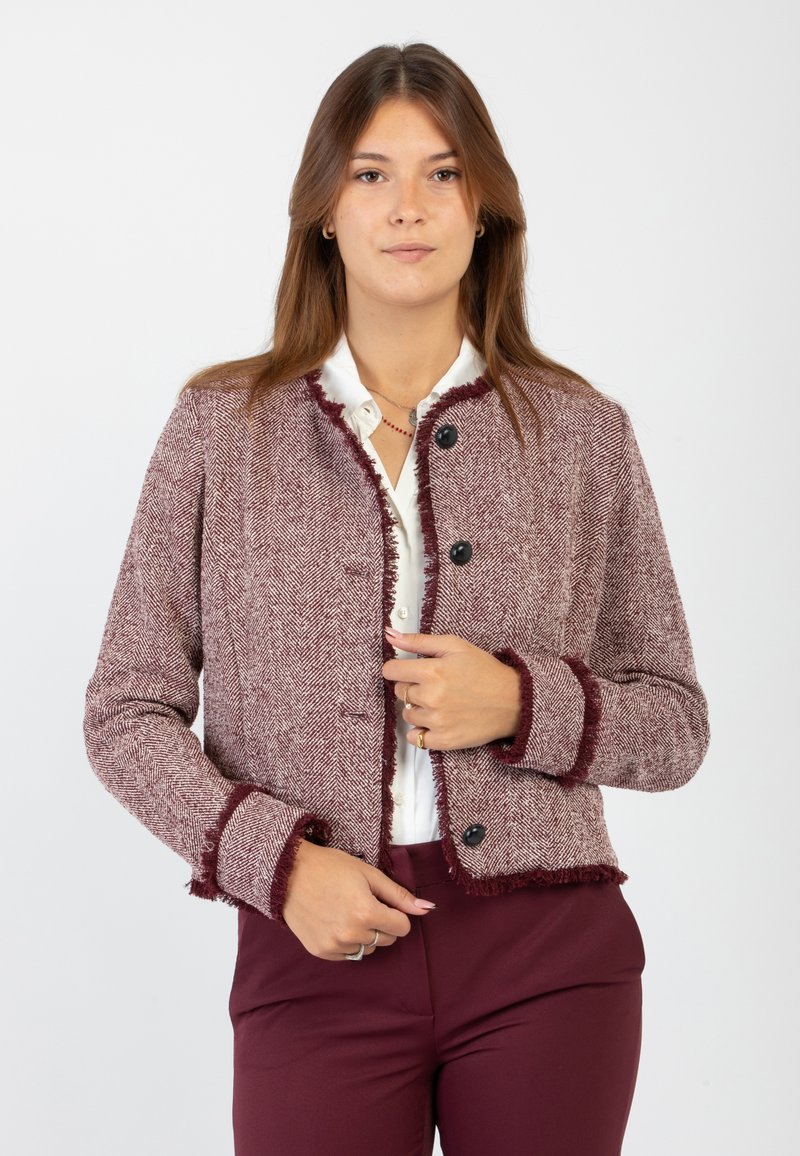 Giacca herringbone bordeaux con bordi sfilacciati, bottoni neri e maniche lunghe. Abbinata a una camicia bianca e pantaloni scuri.