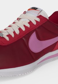 Κόκκινο και λευκό αθλητικό παπούτσι Nike με ροζ λογότυπο Swoosh, λευκά κορδόνια και ραβδωτή λευκή σόλα.