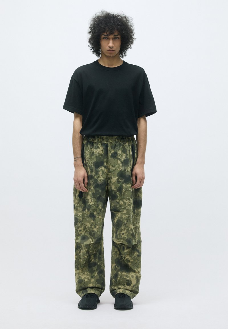 Zwarte oversized t-shirt gecombineerd met losse, gemarmerde cargobroek in verschillende tinten groen en met textuurpatronen. Zwart schoeisel maakt de look af.