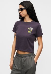 Μωβ κοντομάνικη cropped μπλούζα με κεντήματα λουλουδιών και το λογότυπο της North Face. Υφασμάτινη απαλότητα, φορεμένη με γκρι παντελόνι.