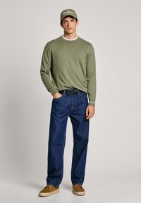 Olivgrön stickad tröja med rundhals, blå denimjeans, bruna skor och en matchande olivgrön keps med vit logotyp. Rakt ben-passform.