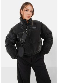 Zwarte pufferjack met een hoge kraag, zijzakken met rits en een afneembare schouderriem. Bevat een logo op de borst.