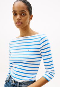 Μπλε και λευκή ριγέ μπλούζα μακριάς μανικιού με bateau λαιμόκοψη και μικρό λογότυπο στο στήθος, συνδυασμένη με τζιν παντελόνι. Λεία υφή υφάσματος.
