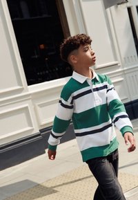 Zeleno-bílé pruhované polo tričko s dlouhým rukávem a námořnicky modrými detaily, spojené s tmavými džíny. Chodí po chodníku poblíž budovy.