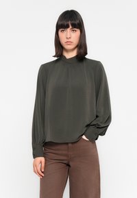 Kvinne med mørkt, rett hår iført en høyhalset, mørkegrønn bluse med lange ermer og brune bukser, stående med den ene hånden i lommen.