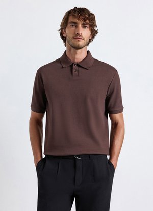 Mann mit lockigem Haar, der ein braunes kurzärmeliges Poloshirt und schwarze Hose trägt, steht mit den Händen in den Taschen vor einem einfarbigen Hintergrund.
