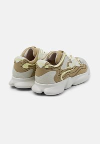Light beige and white sneakers with textured mesh upper, rubber sole, and abstract pattern. Features prominent branding on the heel.
