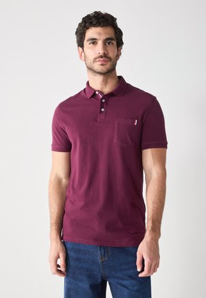 Burgundy polo shirt with short sleeves, three-button placket, and chest pocket featuring a striped accent. Worn with blue jeans.