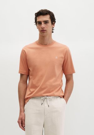 Młody mężczyzna stojący z rękami w kieszeniach, ubrany w łososiowy t-shirt z krótkim rękawem i jasnobeżowe spodnie ze ściągaczem na tle jednolitego tła.