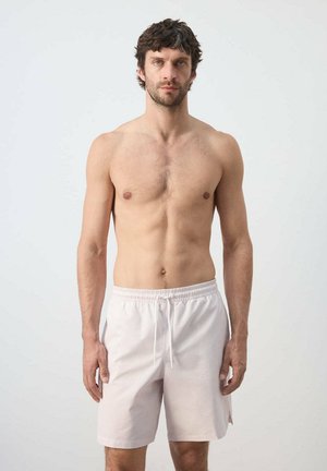 Hombre con cabello oscuro corto, pecho descubierto, vistiendo pantalones cortos claros con cordón, de pie frente a un fondo blanco liso.