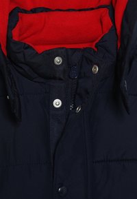 Marinblå pufferjacka med krage av röd fleece. Har tryckknappar och en dragkedja framtill. Mjukt material och vadderad design.