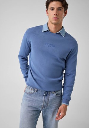 Jeune homme portant un pull bleu à col rond avec le logo « GUESS » sur une chemise en jean à col et un jean bleu clair, debout devant un fond uni.