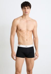 Schwarze Boxer-Shorts aus Stretchmaterial mit einem weißen Bund, auf dem die Marke "BOSS" wiederholt ist. Modell zeigt eine Frontalansicht.