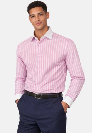 Homme portant une chemise à rayures verticales roses et blanches avec col et poignets blancs, pantalon bleu marine et ceinture noire, les mains dans les poches.