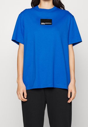Personne portant un t-shirt à manches courtes bleu vif avec un logo carré noir indiquant « KARL LAGERFELD JEANS » sur la poitrine, associé à un pantalon noir.