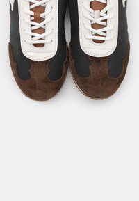 Paire de baskets noires et marron avec des lacets blancs et des détails en cuir, vues de dessus sur fond blanc.