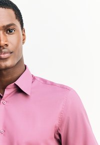 Jasnoróżowa koszula zapinana na guziki z teksturowanym wykończeniem, wyposażona w klasyczny kołnierzyk i standardowe guziki, podkreślona czystymi liniami i dopasowanym krojem.