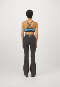 Kvinna som står vänd bort med en blå sportbh med korsade axelband och högmidjade mörka ribbade utsvängda byxor på en vit bakgrund.