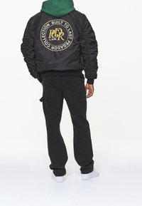 Giacca bomber nera con maniche arricciate, caratterizzata da un grande logo rotondo sul retro in oro e nero. Abbinata a jeans neri e sneakers bianche.