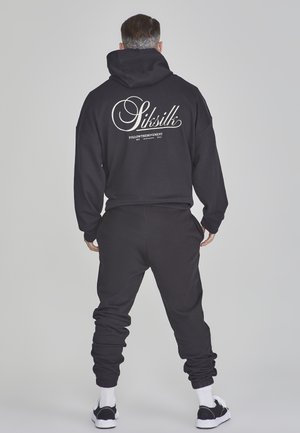 Sudadera con capucha negra y pantalones de chándal hechos de una tela suave, con un gran logo blanco de la marca en la parte posterior. Los puños son de canalé para un ajuste ceñido.