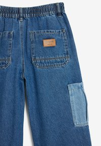 Pantalones de mezclilla con cintura elástica, que presentan un parche de cuero marrón, dos bolsillos traseros y un bolsillo lateral de un azul claro contrastante.