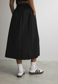 Osoba ubrana w czarną plisowaną spódnicę midi z rękami w kieszeniach, w połączeniu z białymi skarpetkami do kostek i białymi sneakersami Adidas w czarne paski.