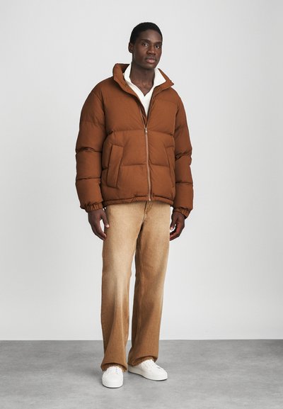 Brun pufferjakke med høj krave, lynlås og sidelommer. Båret over en cremefarvet skjorte med løse beige bukser og hvide sneakers.
