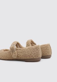 Tan fuzzy slip-on shoes with two adjustable straps, rounded toe, and a flat rubber sole. Textured fabric adds visual interest.