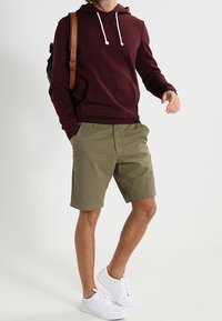 Uomo che indossa una felpa maroon, pantaloni corti verde oliva, sneakers bianche e porta uno zaino con tracolla marrone su una spalla.