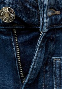 Jeans de denim oscuro con un botón plateado, una cremallera metálica con dientes visibles y costuras de contraste a lo largo de los bordes.