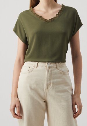 Femme portant un haut texturé vert olive à col en V avec bordure en dentelle festonnée et un pantalon taille haute couleur écru sur fond uni.