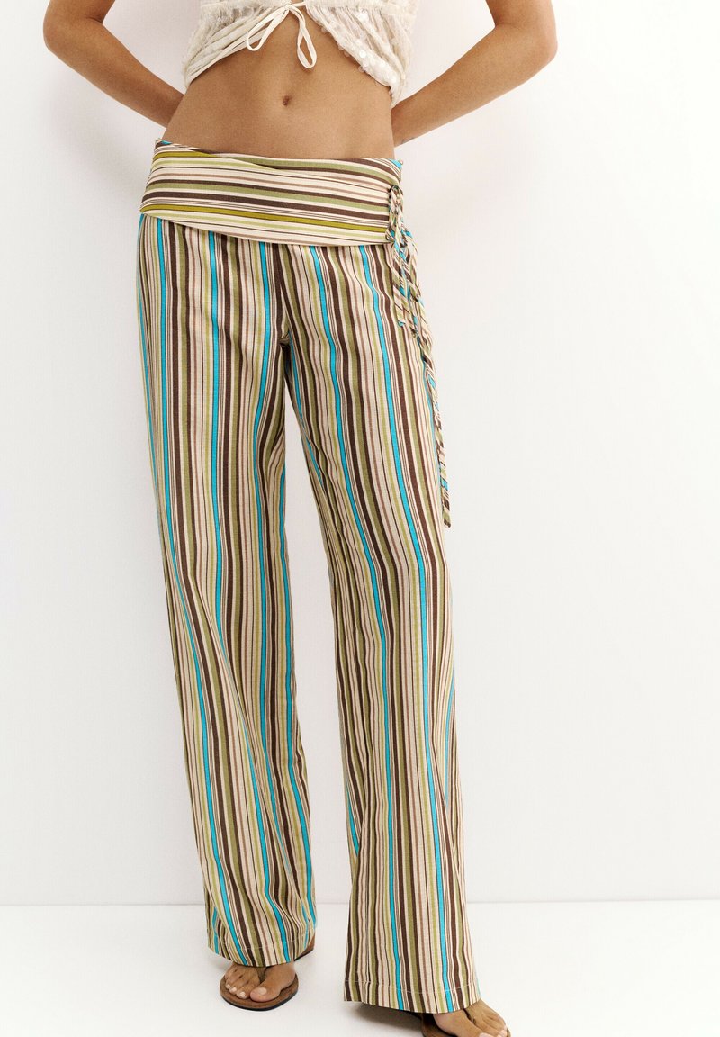 Woman wearing wide-leg pants with vertical stripes in beige, brown, blue, and green, paired with a tied crop top and sandals.