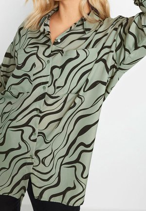 Mujer con una camisa verde abotonada con un patrón abstracto de líneas onduladas negras, con el brazo levantado y el cabello rubio y ondulado.