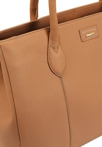 Sac fourre-tout en cuir tan avec deux poignées, présentant des accents cousus et une petite plaque logo en métal. Texture lisse, forme structurée.