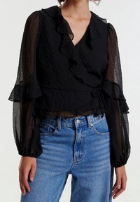 Blouse noire texturée avec un col et des manches volantés. Nouée à la taille. Associée à un jean taille haute bleu clair.
