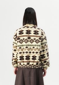 Persoon met steil zwart haar die een crèmekleurige fleecejack draagt met donkerbruine en groene geometrische patronen, staande tegen een effen achtergrond.