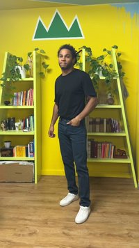 Musta t-paita, tummat farkut ja valkoiset lenkkarit; seisoo keltaisen seinän edessä, jossa vihreä vuoristografiikka ja vihreäkirjahylly.