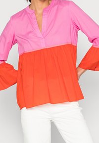 Blusa a blocchi di colore con una parte superiore rosa e una parte inferiore arancione. Stile tunica con una leggera arricciatura e maniche a campana.