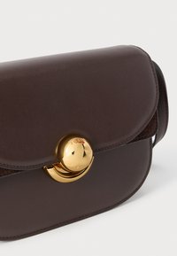 Dunkelbraune Leder-Umhängetasche mit abgerundeten Kanten und einem glänzenden, goldfarbenen, runden Verschluss, der mit Markentext graviert ist.