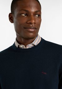 Marineblauer strukturiertes Strickpullover mit einem gerippten Kragen, über einem gestreiften Hemd mit Kragen getragen. Verfügt über ein kleines gesticktes Logo auf der Brust.