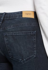 Jeans de mezclilla azul oscuro con un acabado texturizado. Incluye un parche de logo de cuero marrón y costuras en contraste en los bolsillos traseros.