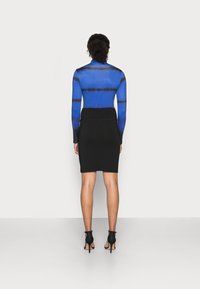 Femme aux cheveux bouclés se tenant face à un mur, portant un haut à manches longues bleu et noir en tie-dye, une jupe crayon noire et des sandales à talons noirs.