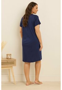 Kvinna stående barfota i en knälång marinblå kortärmad klänning, vänd mot en beige vägg nära ett träbord med ett ljus.