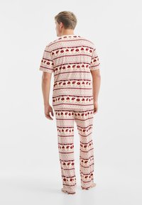 Heren pyjamastel in zacht, lichtgewicht katoen. Heeft een crèmekleurige basis met rode en gouden patroonstripes. Losse pasvorm, korte mouwen en lange broeken.