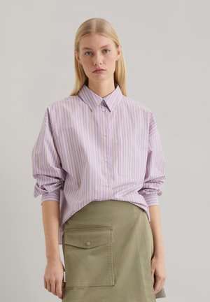 Chemise rayée mauve clair à col boutonné, avec des manches retroussées et une poche sur la poitrine, assortie à une jupe verte olive.