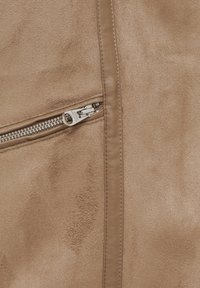 Gros plan sur un tissu en daim beige avec une fermeture éclair métallique horizontale et une bordure en cuir cousue en vertical.