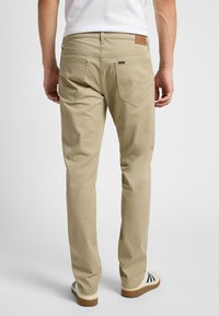 Pantalones caqui de corte recto, con bolsillos traseros y un parche de cuero encima del bolsillo derecho. El material parece suave; diseño casual.