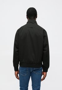 Sort bomberjakke lavet af glat stof med ståkrave, ribstrikkede manchetter og et diskret brandmærke bagpå. Kombineret med blå jeans.